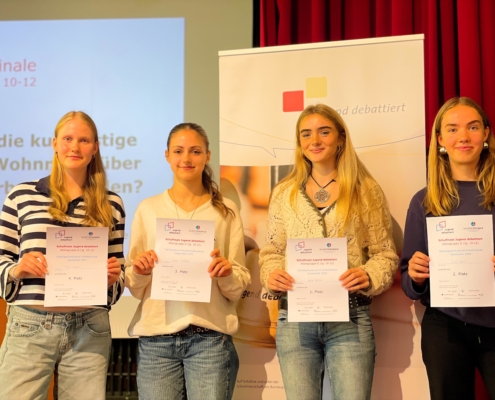 Vier Schülerinnen stehen auf der Bühne der Aula und halten ihre Urkunden in die Kamera