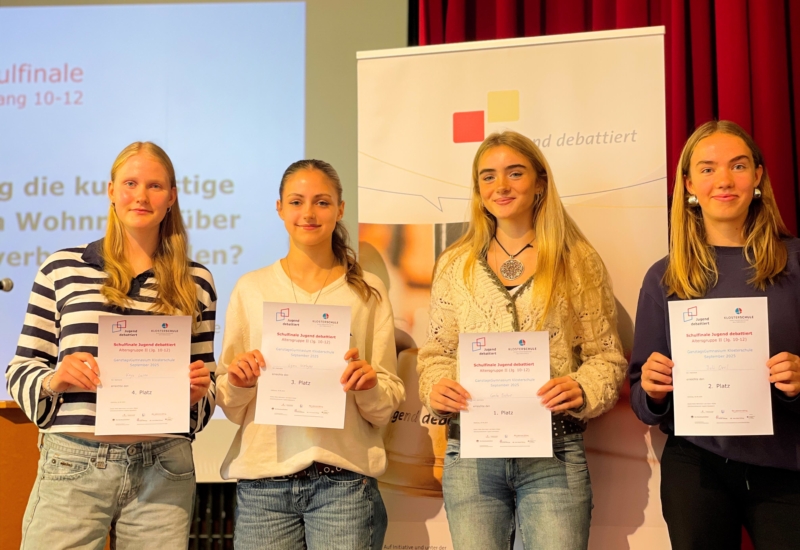 Vier Schülerinnen stehen auf der Bühne der Aula und halten ihre Urkunden in die Kamera