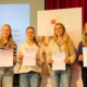 Vier Schülerinnen stehen auf der Bühne der Aula und halten ihre Urkunden in die Kamera