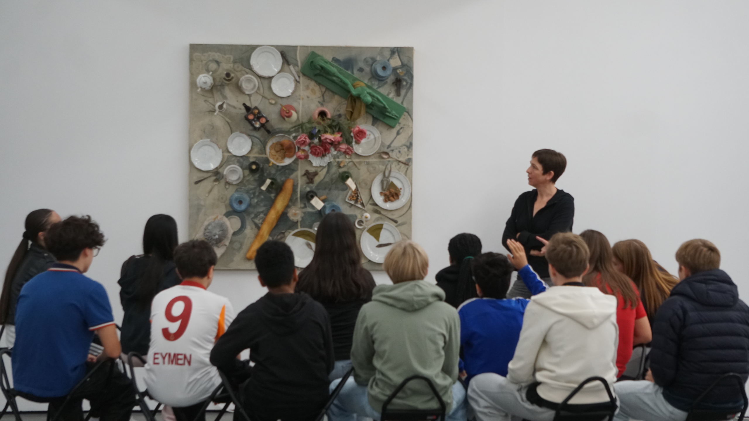 Schülergruppe von hinten sitzend vor einer an der Wand hängenden Collage; Frau betrachtet die Collage, Schüler zeigen auf