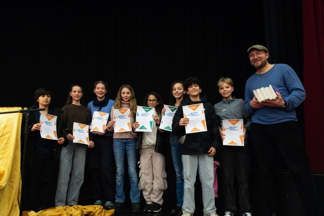 Die Teilnehmer:innen des Vorlesewettbewerbs stehen mit ihrem Lehrer auf der Bühne und halten Urkunden in die Kamera