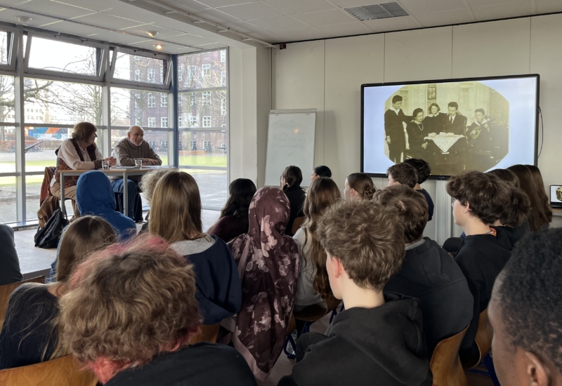 Bernhard Esser und Barbara Brix sitzen auf einer Bühne und sprechen zu Schülern des 9. Jahrgangs