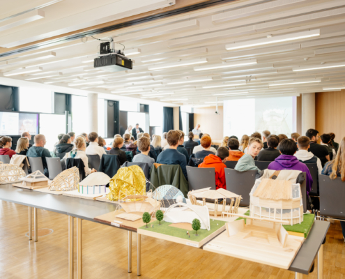 Blick auf Architekturmodelle und Schüler von hinten. Vorne findet auf einer Bühne die Preisverleihung statt.