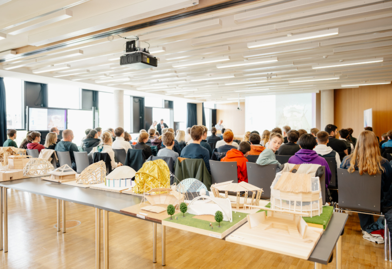 Blick auf Architekturmodelle und Schüler von hinten. Vorne findet auf einer Bühne die Preisverleihung statt.