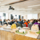 Blick auf Architekturmodelle und Schüler von hinten. Vorne findet auf einer Bühne die Preisverleihung statt.